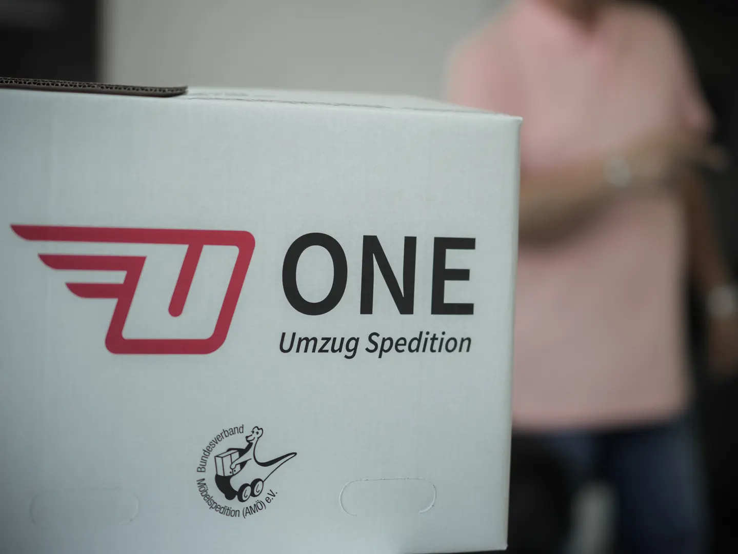 Karton mit der Aufschrift „One Umzugsspedition GmbH“ und dem Logo mit einem stilisierten U und Flügeln. Ein kleineres Symbol zeigt einen Schuh und einen Mann, der Umzugskartons trägt. Im Hintergrund verleiht eine verschwommene Person in einem rosa Hemd der Szene Tiefe.