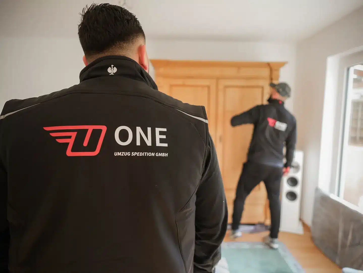 Zwei Umzugshelfer in schwarzen Jacken mit dem ONE-Logo sorgen für Umzugsqualität bei der Arbeit in einem Raum. Einer greift nach einem Schrank, der andere steht mit dem Rücken zur Kamera im Vordergrund. Der Raum ist mit Holzboden und einem großen Fenster ausgestattet.
